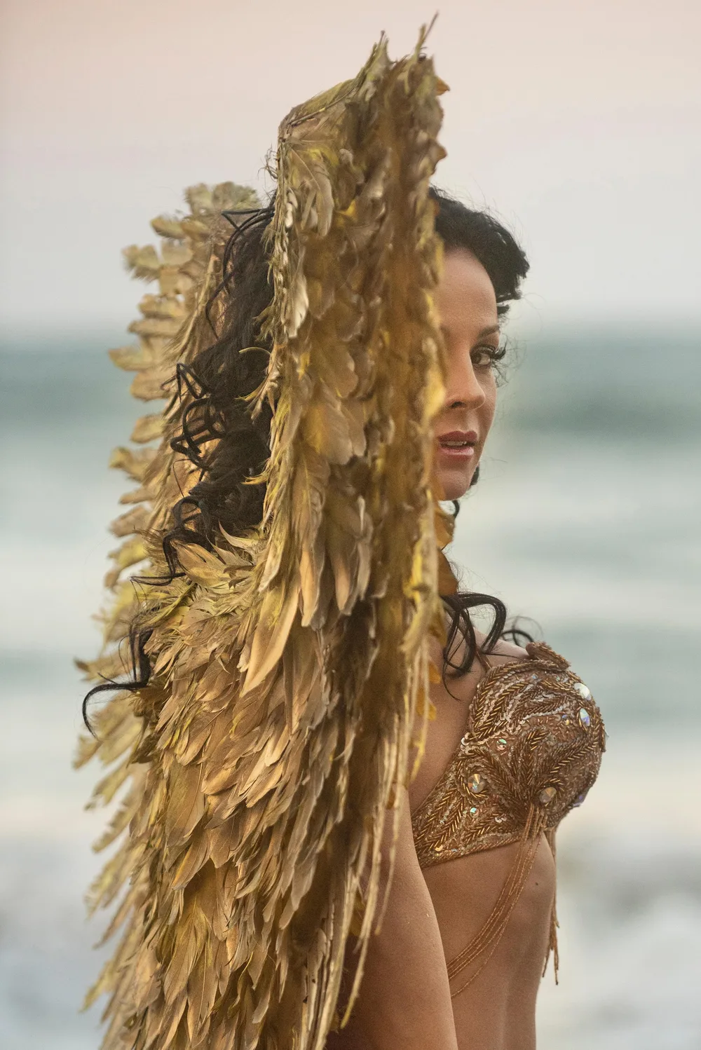 Verónica Pavón beside the sea, wearing a golden costume and holding a feather fan.